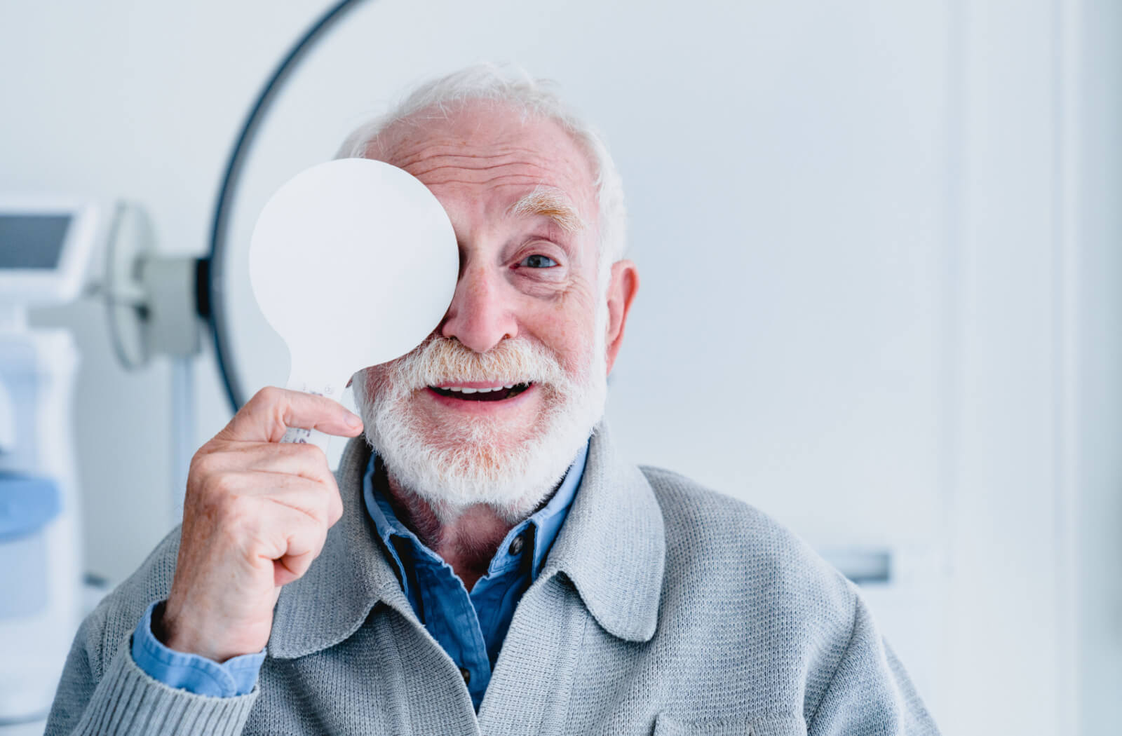 A happy senior male is covering his right eyed during a vision acuity exam in an optician's clinic.