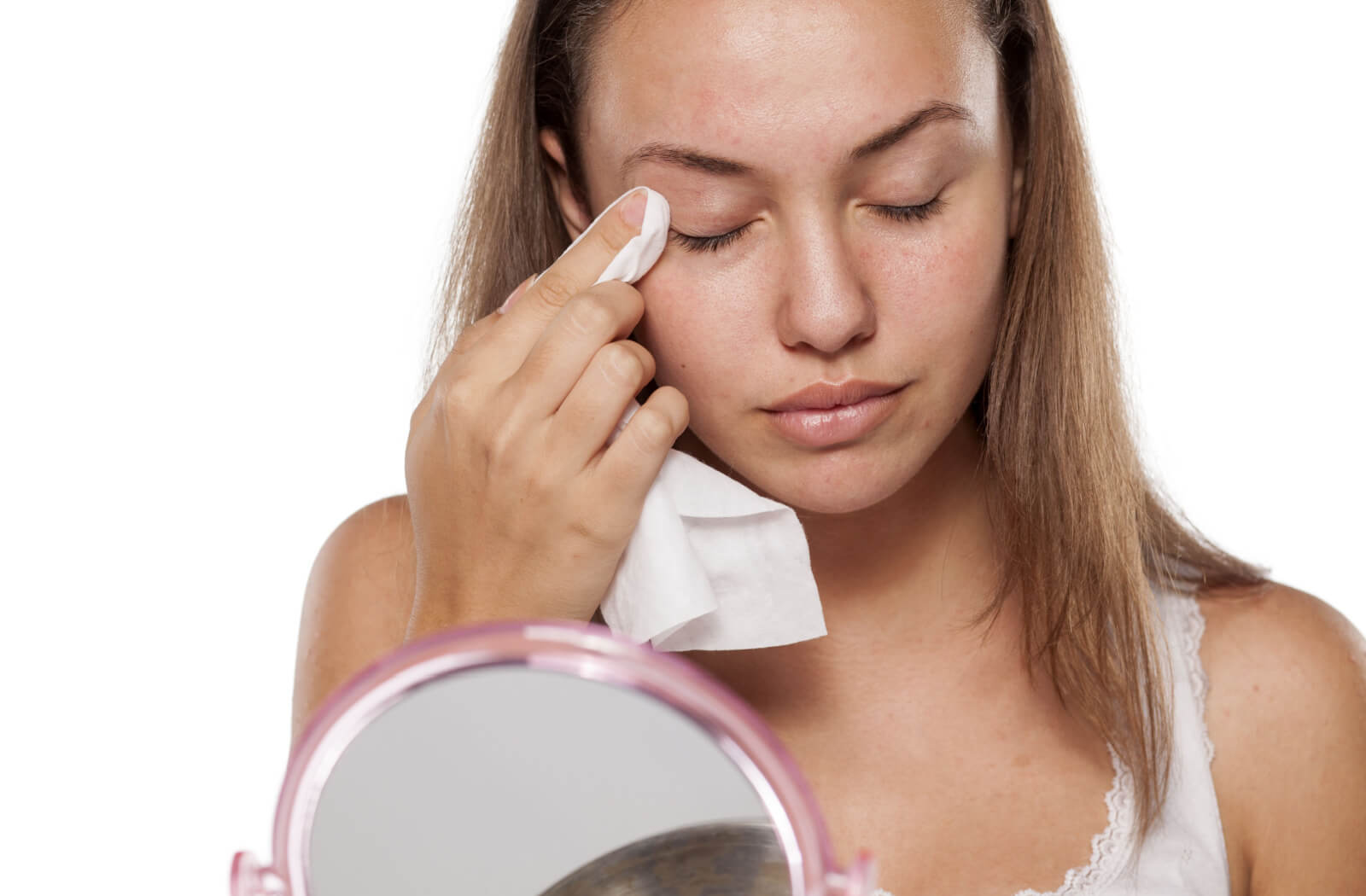 A young woman is taking off her makeup and cleansing her eyelids with wet wipes while sitting in front of a mirror.