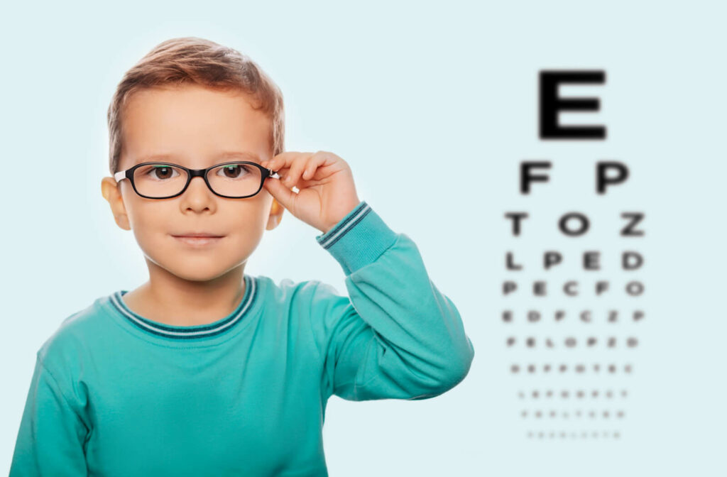 A child looking at the camera, adjusting his eyeglasses with an eye chart in the background