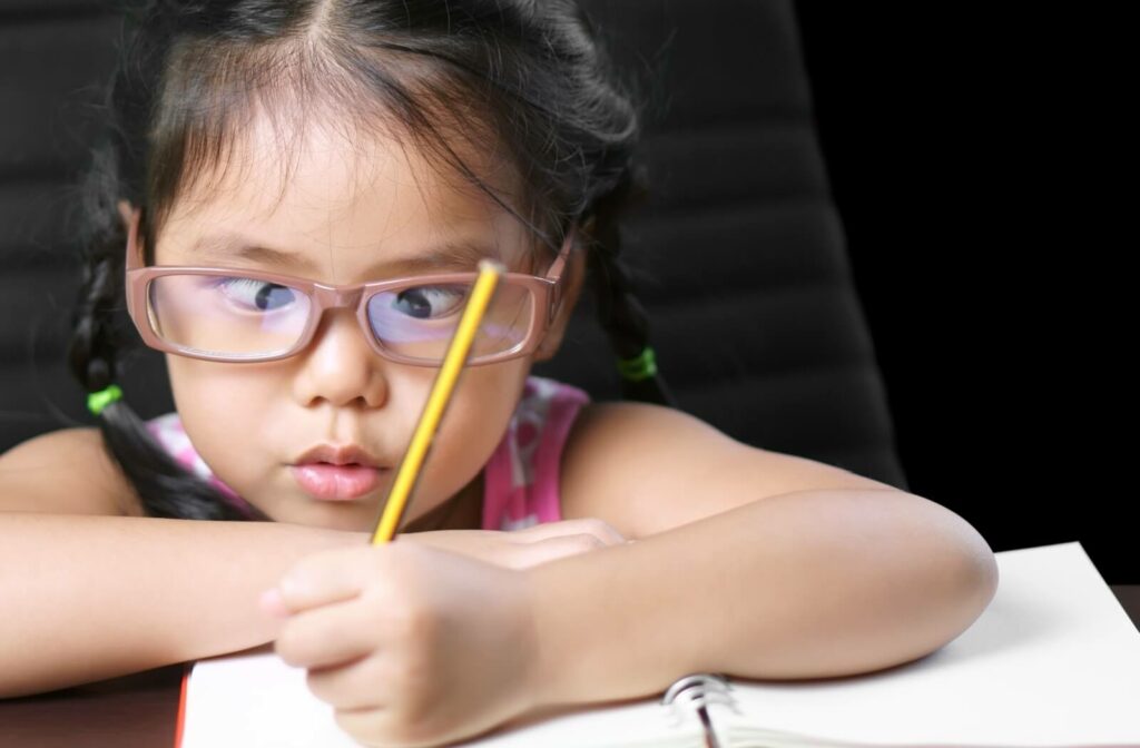 A young girl with a lazy eye uses glasses to help write in a notebook. Her arms rest on the table while she holds a pencil