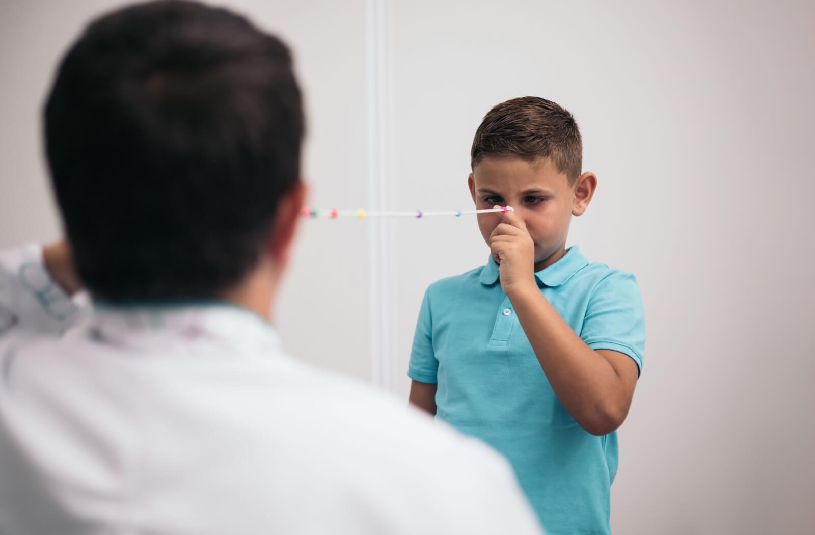 A young boy in a blue shirt holds a Brock String to his eye, focusing on the beads, while an optometrist holds the other end