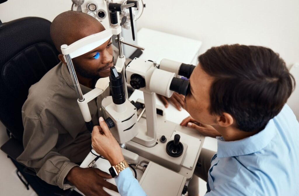 A patient sits in the examination chair at an optometrist's office. The optometrist sits across from the patient, examining the patient's eye through a slit lamp.