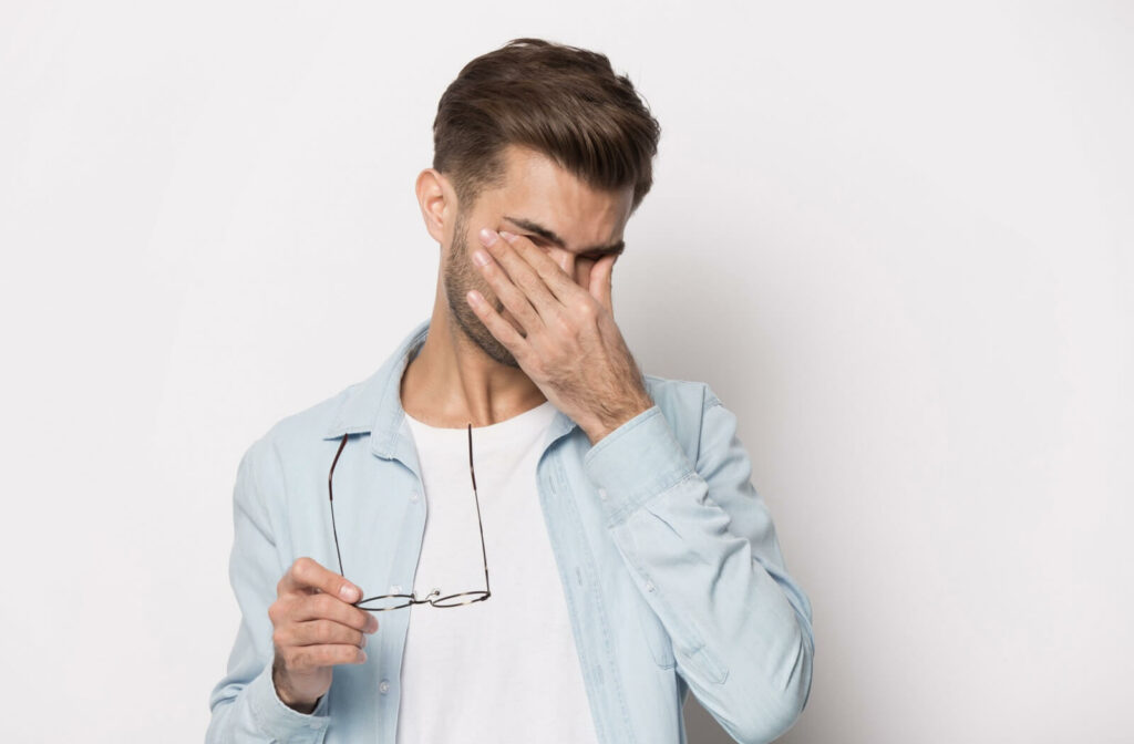 A young adult in front of a white background taking off their glasses to rub their eyes due to dry eyes and discomfort.