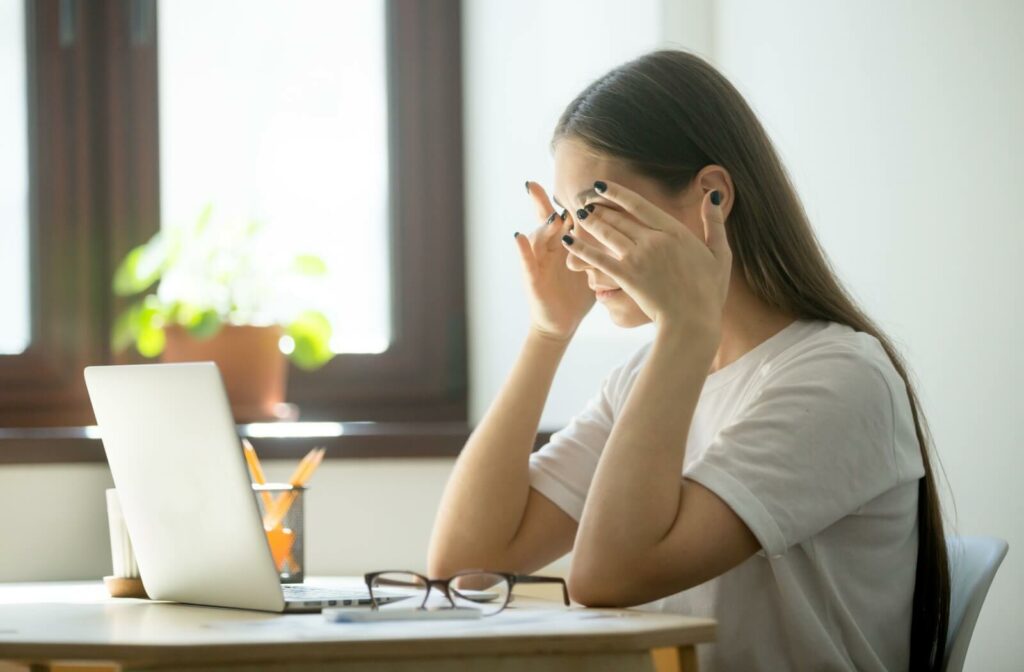 A young adult working at their desk, taking a break and rubbing their eyes due to headaches and dry eye disease.