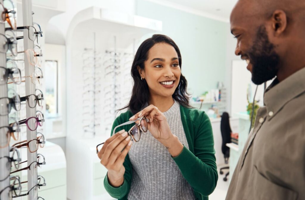 A smiling person in a green sweater shows a pair of eyeglasses to a person inside an optical store.