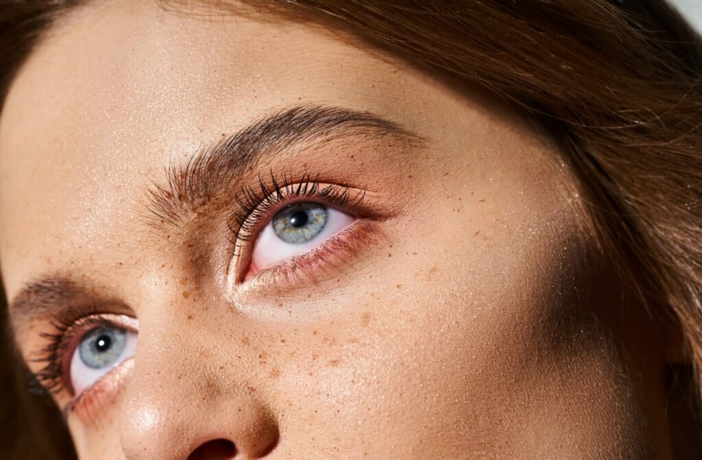 Close-up side view of a person with light blue eyes and freckles, showing detailed iris texture, eyelashes, and natural skin tone under bright, directional lighting that emphasizes eye color and light exposure.