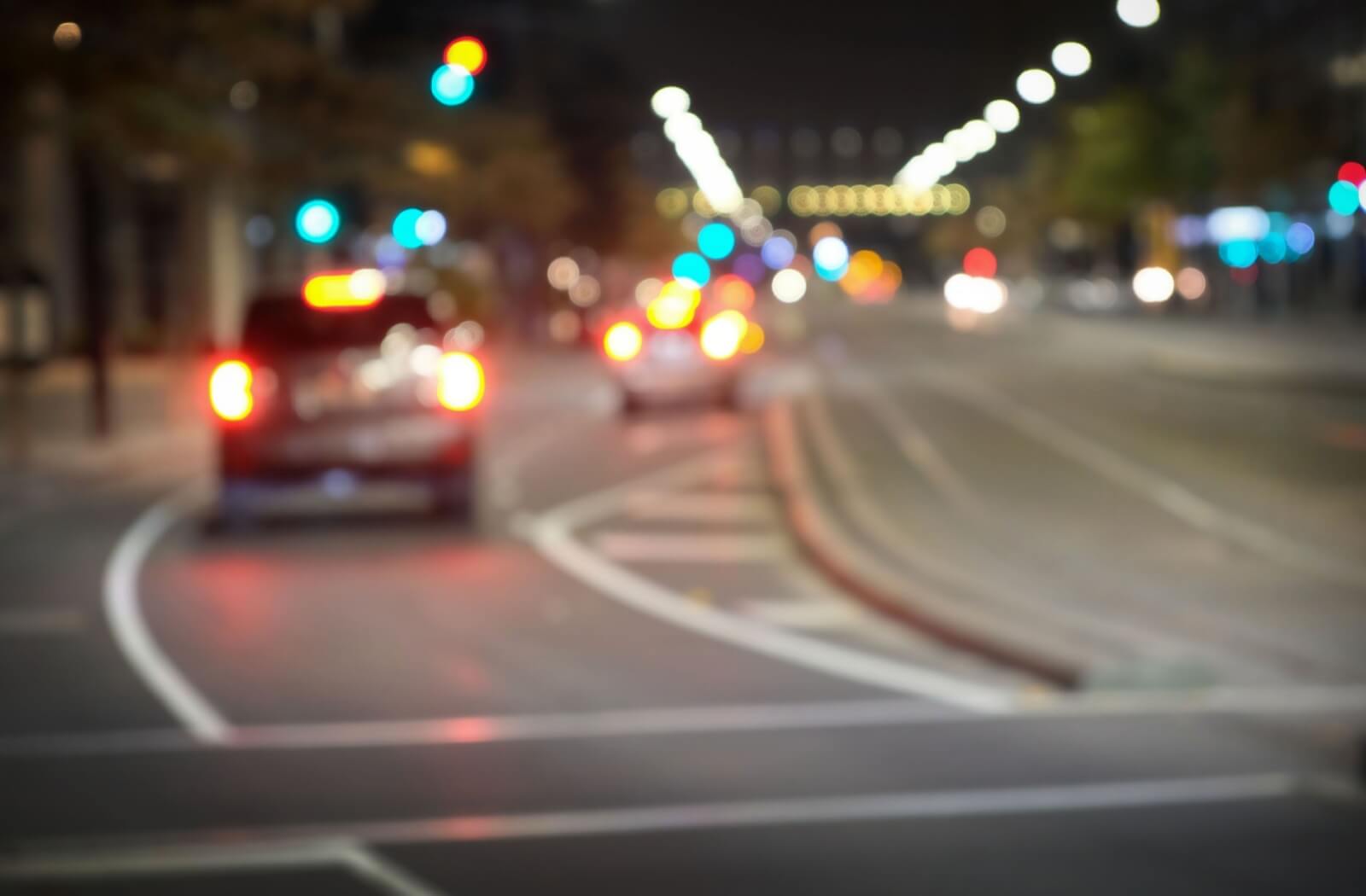 Blurred nighttime street scene with out-of-focus headlights and traffic lights, representing blurry vision, poor night vision, and visual disturbances such as glare and halos while driving at night.