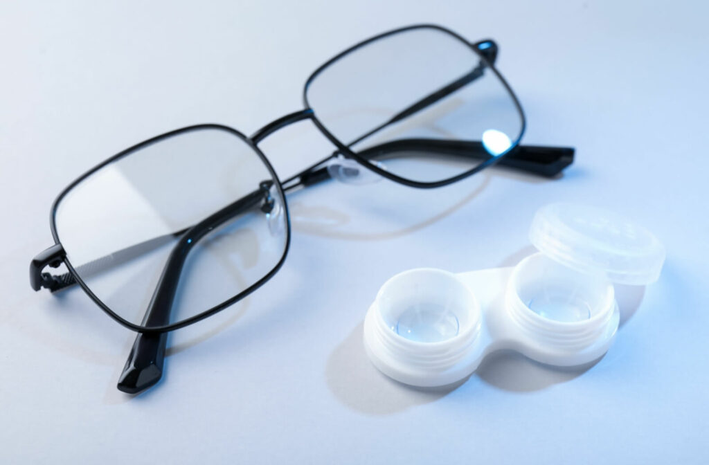 A pair of black-framed eyeglasses next to an open white contact lens case on a light blue surface.