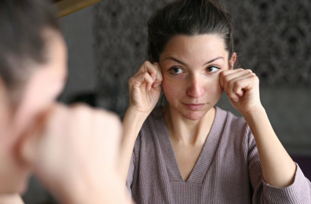 An individual looking into a mirror and gently pulling down their lower eyelids to examine their eyes for redness or irritation.
