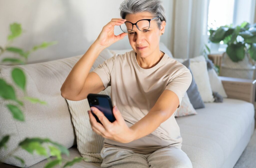 A middle-aged woman with grey hair lifting her glasses and squinting at her smartphone while sitting on a couch, illustrating symptoms of presbyopia or declining vision.
