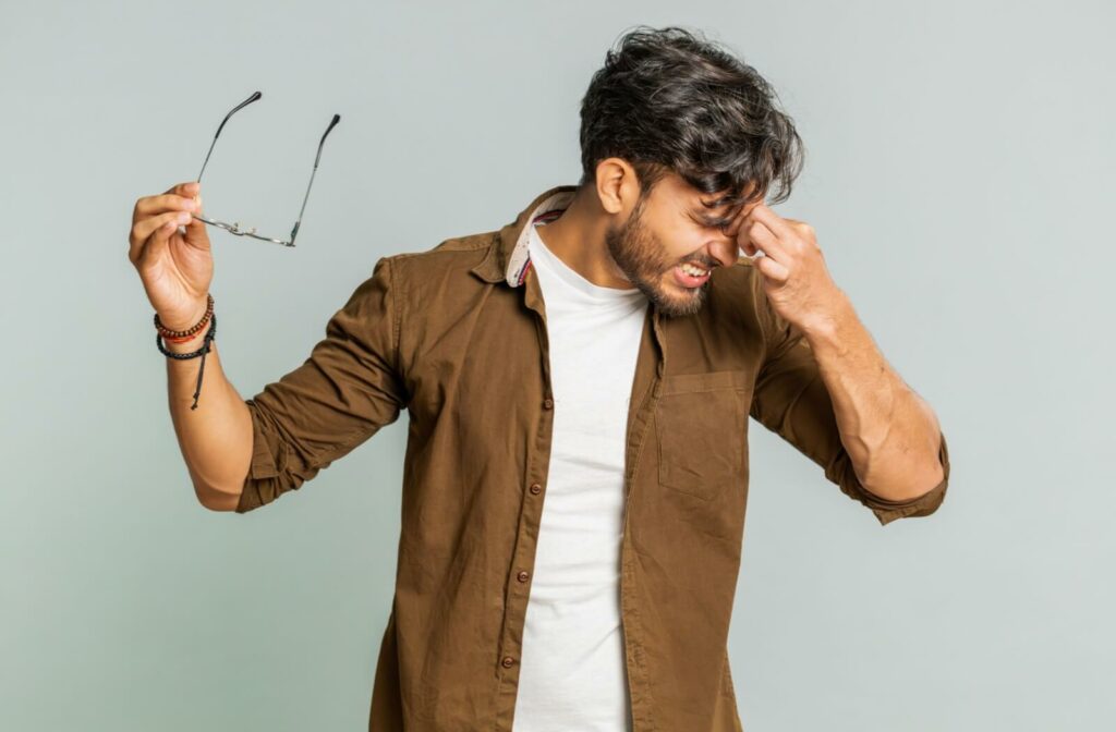 A man holding his glasses in one hand while pinching the bridge of his nose and grimacing, illustrating digital eye strain and vision-related headaches.